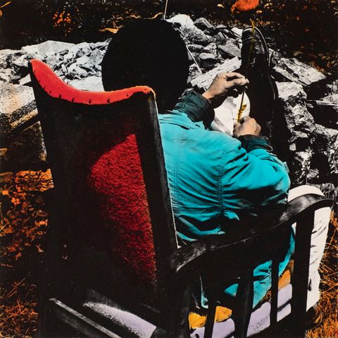 Hand-coloured black and white photograph. A man sits facing away from us in a high-backed armchair. The back of the chair is vivid red, and the jacket he wears is turquoise. One foot is on the ground and the other foot is propped up on a rock. The person holds something slender, perhaps a twig, in his peach-toned hands.