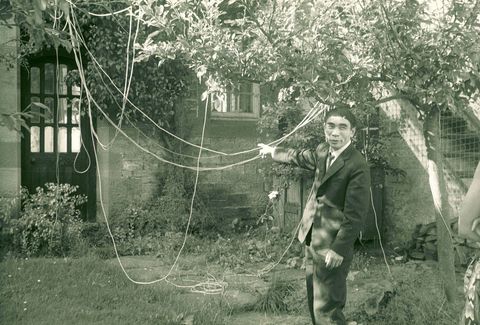 Black and white photograph. A man wearing a suit smiles at someone to our right as he points to strings hanging from trees in an overgrown courtyard. He has a bowl haircut, thick brows, and dimples.
