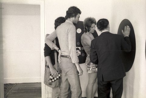 Black and white photograph. Four people stand at a black disc, about half the height of the people, on a gallery wall. A person facing away from us, wearing a suit, gestures towards the disc.